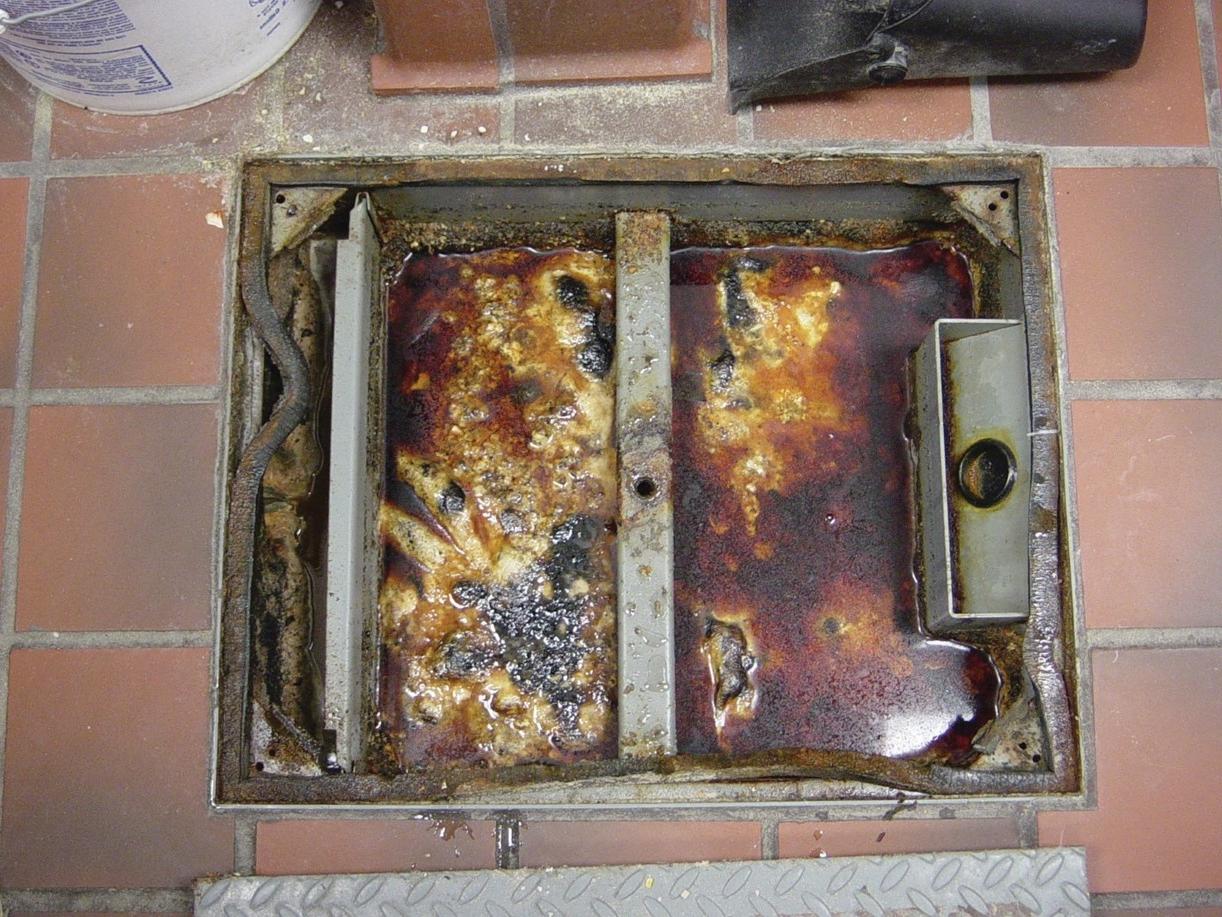  A picture of the floor with a grease trap open in the middle of the floor. The grease trap is a square hole in the floor. This square floor is filled with solidified grease.